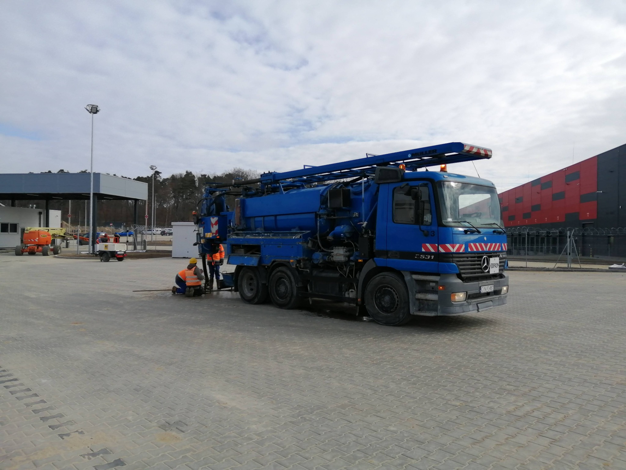 Niebieski samochód asenizacyjny Mercedes-Benz Actros 2531 z długim ramieniem ssącym, dwóch pracowników w kamizelkach odblaskowych przy studzience kanalizacyjnej na placu z kostki brukowej.