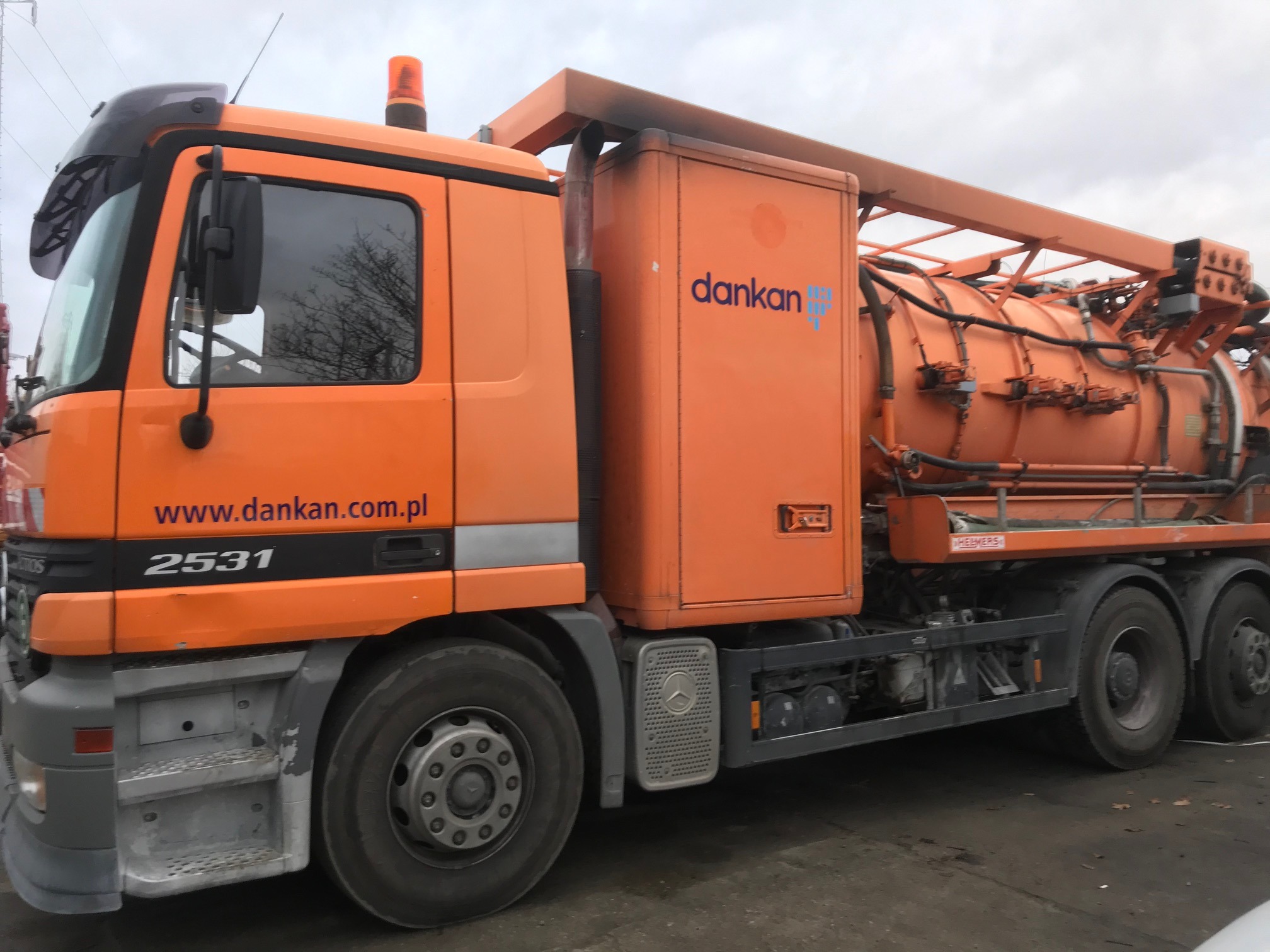 Pomarańczowa ciężarówka do udrażniania rur Dankana z Sopotu, z widocznym logo firmy i numerem 2531, zaparkowana na szarym asfalcie pod pochmurnym niebem.