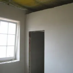 Wnętrze pokoju w stanie surowym po nałożeniu jasnoszarego tynku cementowo-wapiennego na ścianach, widoczne okno z białymi ramami i niedokończone obramowanie drzwi.