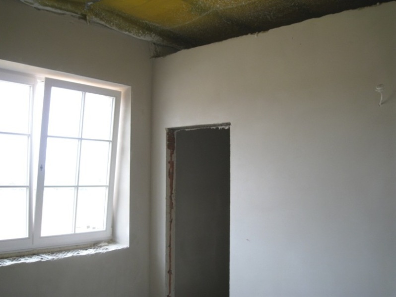 Wnętrze pokoju w stanie surowym po nałożeniu jasnoszarego tynku cementowo-wapiennego na ścianach, widoczne okno z białymi ramami i niedokończone obramowanie drzwi.