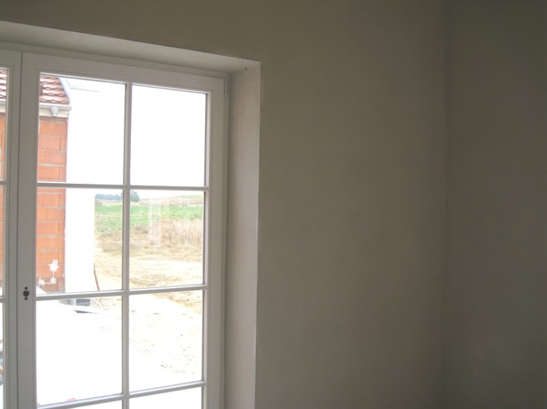 Gładka, jasna ściana po nałożeniu tynku, widoczna obok białego okna z widokiem na pole i budynek w trakcie budowy.