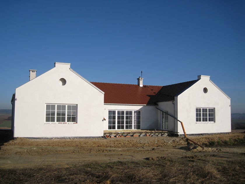 Biały dom z tynkiem cementowo-wapiennym w trakcie prac wykończeniowych, widoczne rusztowania i przygotowany teren wokół budynku.