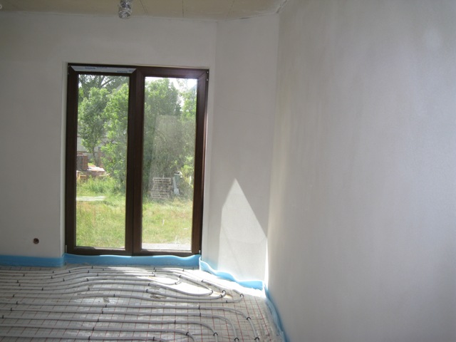 Świeżo otynkowane ściany w pokoju z oknem na ogród i zamontowanym ogrzewaniem podłogowym w trakcie prac wykończeniowych.