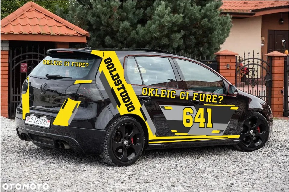Czarny Volkswagen Golf z żółto-szarym oklejeniem reklamowym z napisem 'OKLEIC CI FURE?' i 'GOLDSTUDIO' zaparkowany na żwirowym podjeździe przed domem.