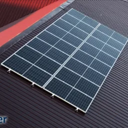 Dwa panele fotowoltaiczne zamontowane na brązowym, blaszanym dachu z widocznym fragmentem czerwonego dachu w tle.