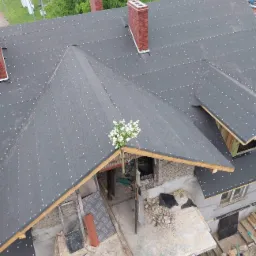 Widok z góry na dach w trakcie pokrywania papą, z kominami z czerwonej cegły i bukietem białych kwiatów wetkniętym w konstrukcję dachu, widoczne elementy więźby dachowej.