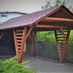 Drewniana wiata garażowa z brązowym dachem, o konstrukcji z ukośnymi belkami i poziomymi szczeblami, przylegająca do garażu z ciemnymi drzwiami, na tle zieleni.