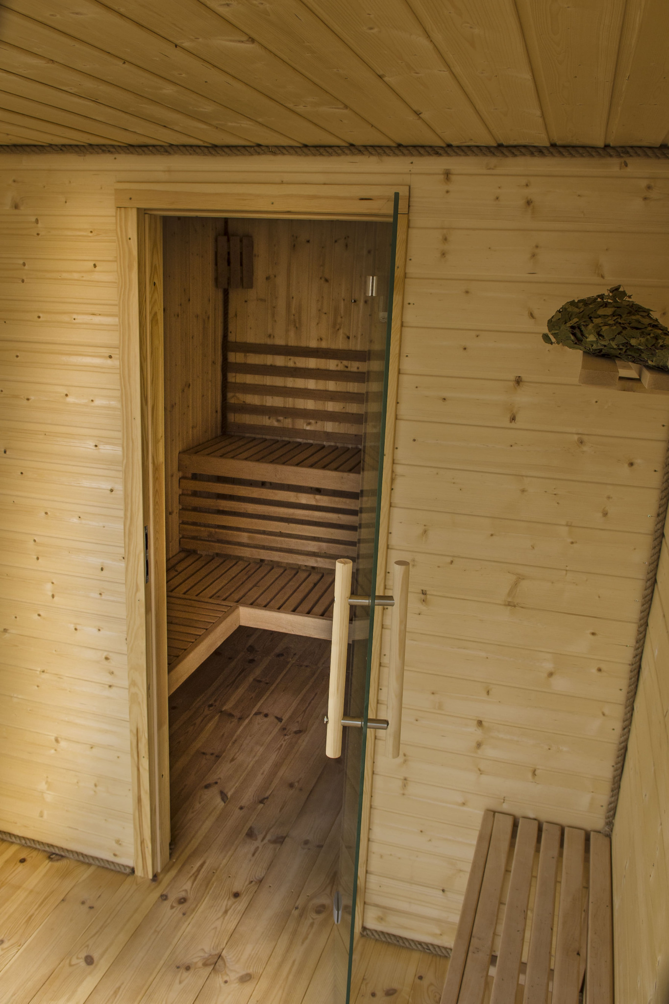 Wnętrze sauny wykończone jasnym drewnem, z przeszklonymi drzwiami i drewnianymi ławkami oraz tradycyjną witką brzozową na ścianie.