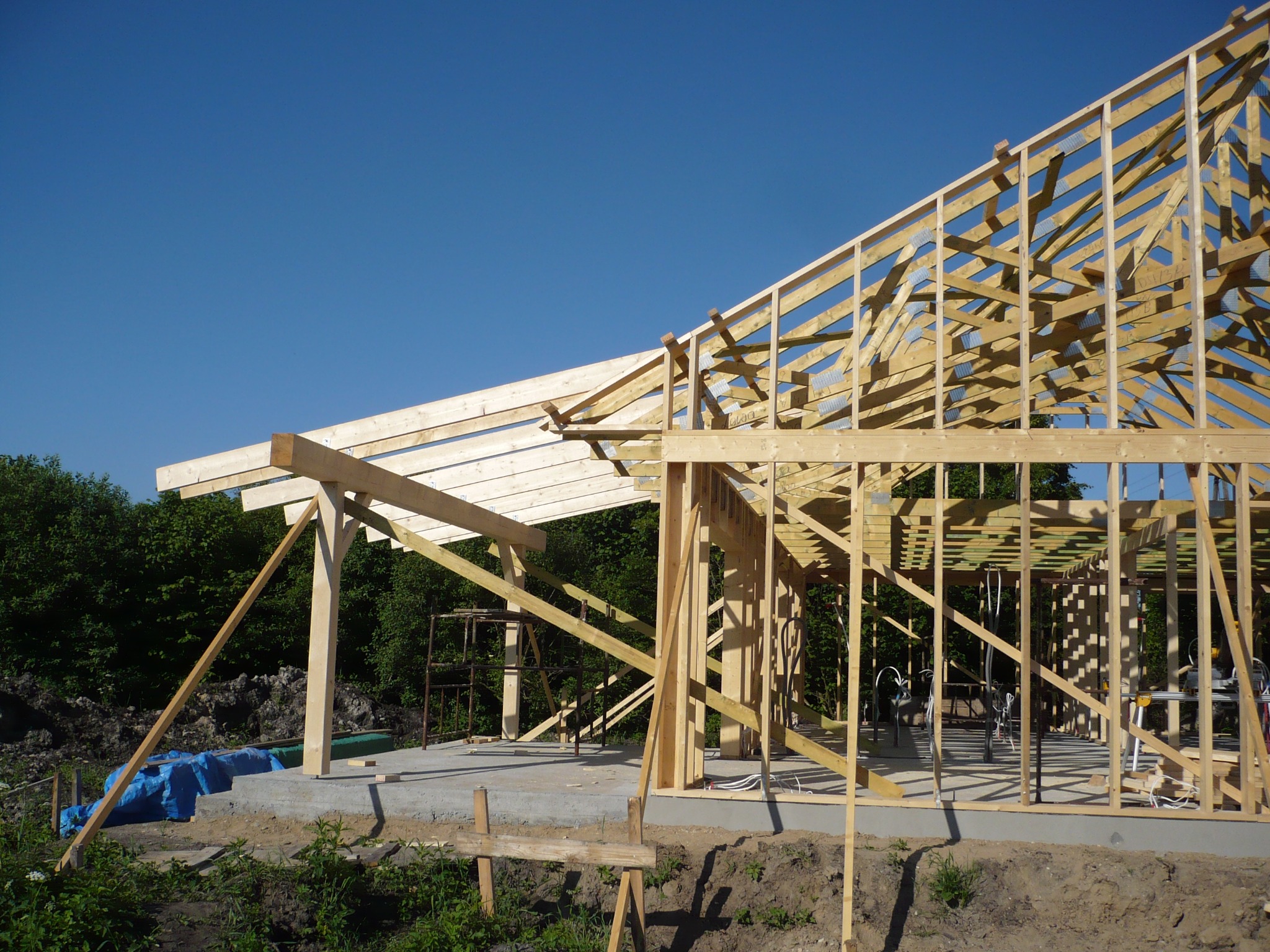 Surowy szkielet drewniany domu w trakcie budowy, widoczne elementy konstrukcyjne pod jasnym, błękitnym niebem, z zielonym tłem drzew.