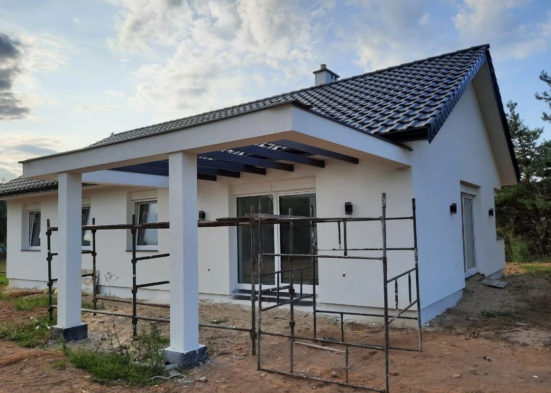 Nowoczesny, parterowy dom z białymi ścianami i ciemną dachówką, zadaszony taras z białymi kolumnami i czarną konstrukcją, widoczne rusztowania, teren wokół domu w trakcie zagospodarowywania.
