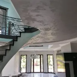 Eleganckie wnętrze w trakcie prac wykończeniowych: sufit pokryty dekoracyjną szarą strukturą przypominającą beton architektoniczny, marmurowe płytki podłogowe odbijające światło, widoczne schody...