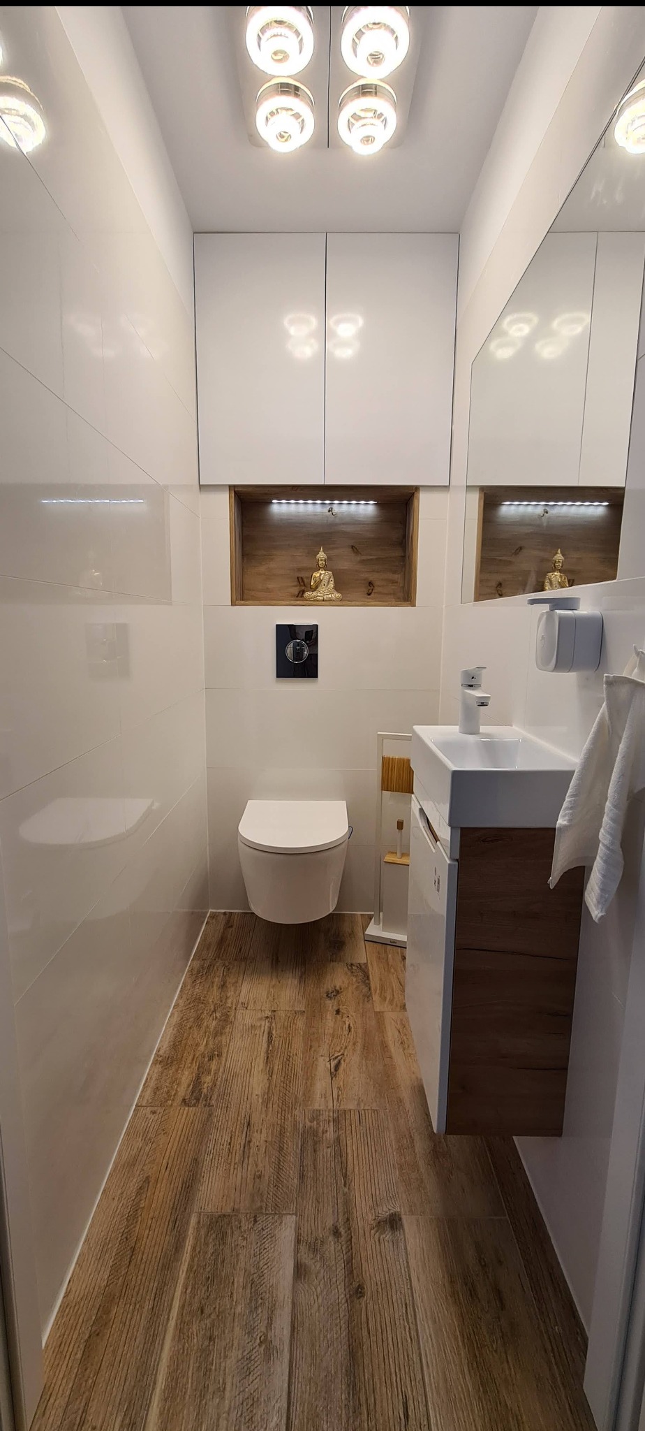 Nowoczesna toaleta z białymi płytkami, drewnopodobną podłogą i szafkami, z dekoracyjną wnęką oświetloną LED z figurką Buddy.