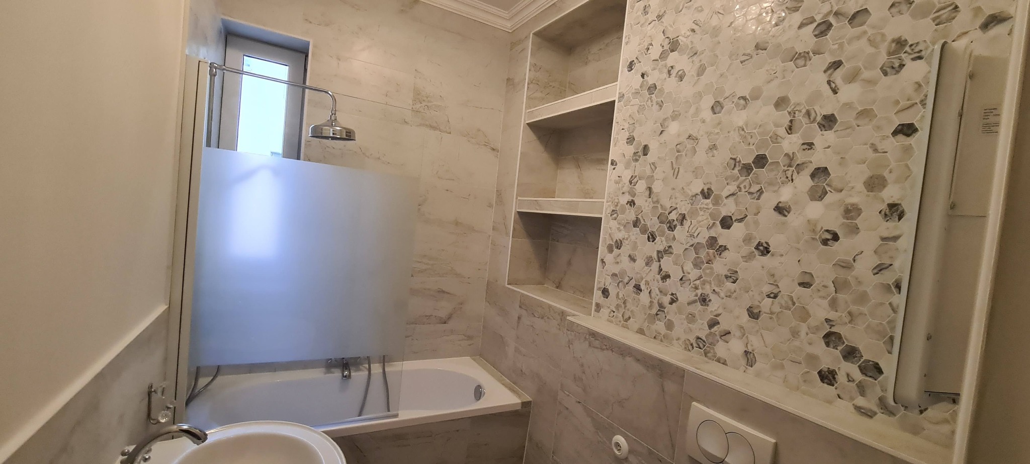 Wykończona łazienka w jasnych barwach z mozaiką heksagonalną na ścianie, kabina prysznicowa z matową szybą, wnęki z półkami i marmurowe płytki.