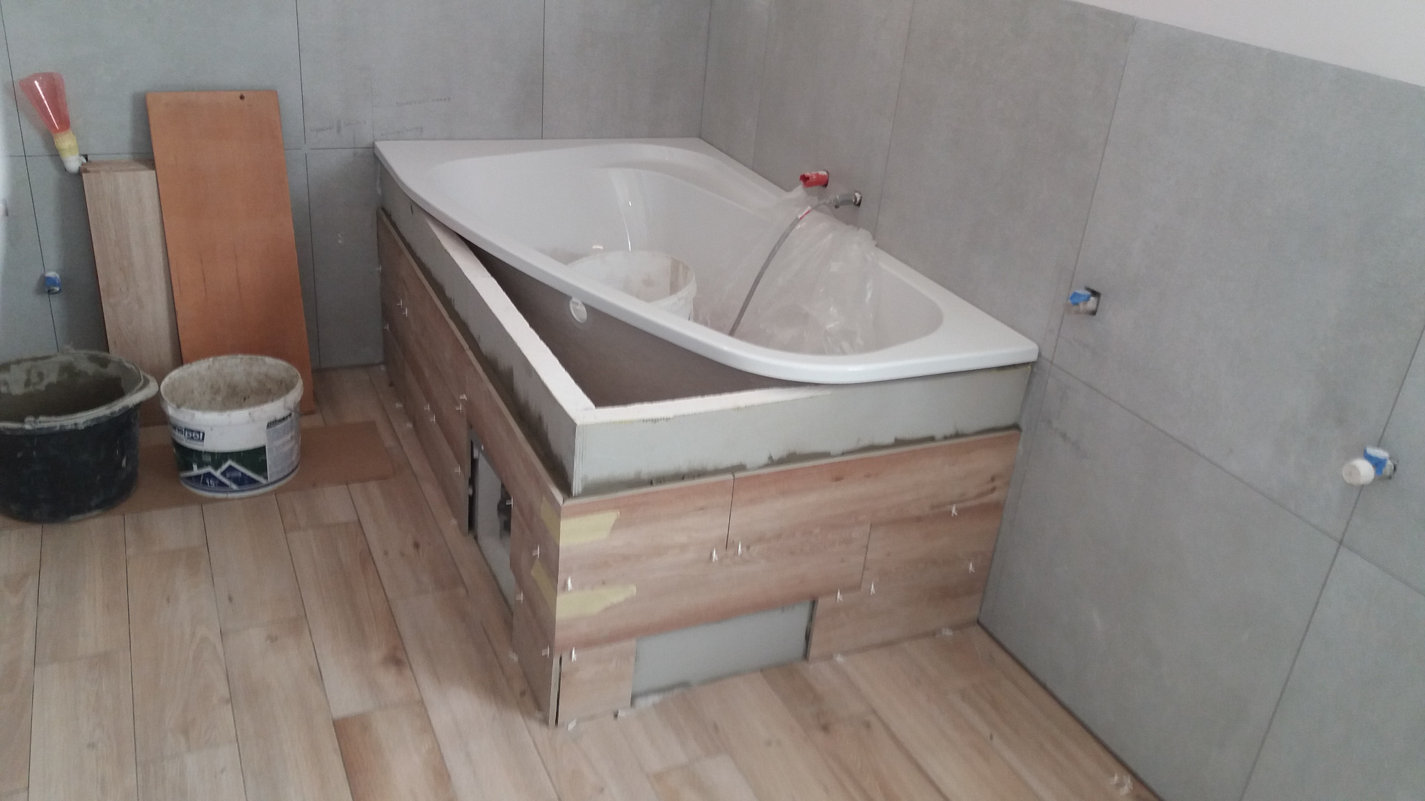 Montaż obudowy wanny z płytek imitujących drewno w narożnej łazience, widoczne narzędzia i materiały budowlane, etap prac wykończeniowych.