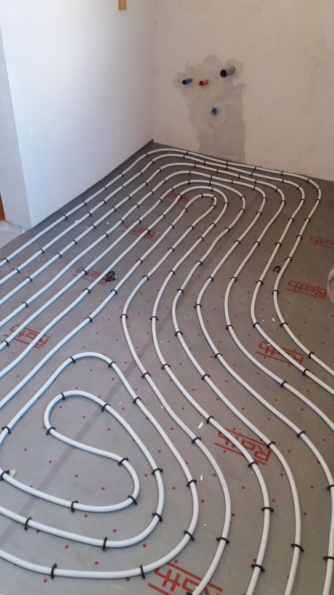 Układanie ogrzewania podłogowego z białych rur na szarej macie izolacyjnej z czerwonym nadrukiem, widoczne czarne mocowania, surowa ściana z instalacją hydrauliczną w tle.