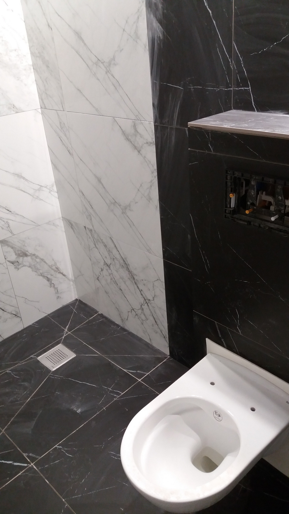 Montaż podwieszanej toalety na tle czarno-białych marmurowych płytek w nowoczesnej łazience, widoczny odpływ podłogowy i elementy instalacji sanitarnej w ścianie.