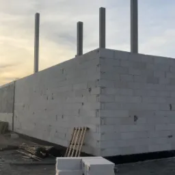 Wznoszone ściany budynku z betonu komórkowego, widoczne cztery betonowe słupy konstrukcyjne, paleta z bloczkami i materiały budowlane na placu budowy.