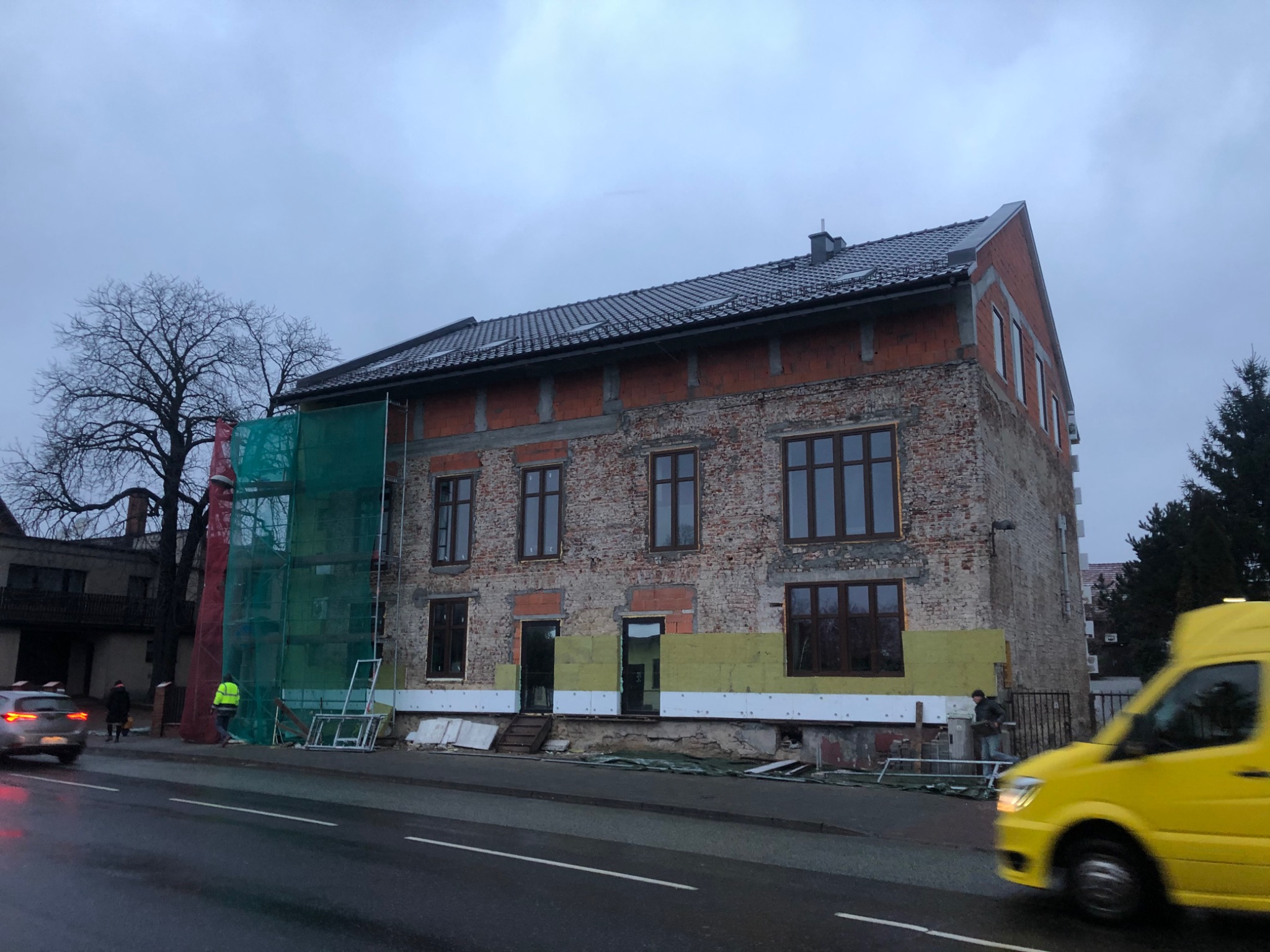 Renowacja ceglanego budynku z częściowo ocieplonymi fundamentami, widoczne rusztowania i pracownicy w odblaskowych kamizelkach, przejeżdżający żółty van.