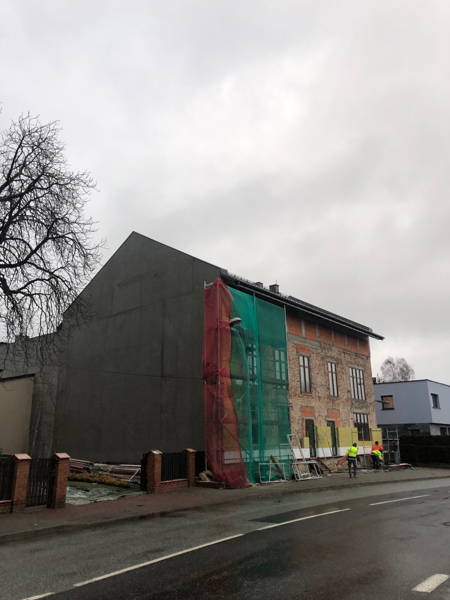 Budynek w trakcie renowacji z częściowo ocieploną i otynkowaną ścianą szczytową, rusztowania z siatką ochronną, oraz pracownicy w kamizelkach odblaskowych przy elewacji z odsłoniętą cegłą.