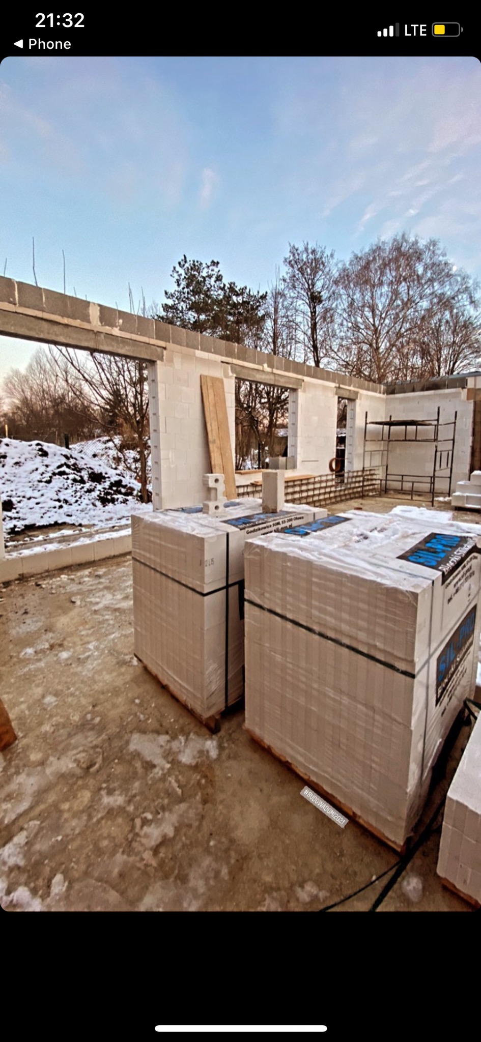 Wznoszenie ścian domu jednorodzinnego z betonu komórkowego; widoczne palety z bloczkami, częściowo wymurowane ściany z otworami okiennymi i resztki śniegu na ziemi.