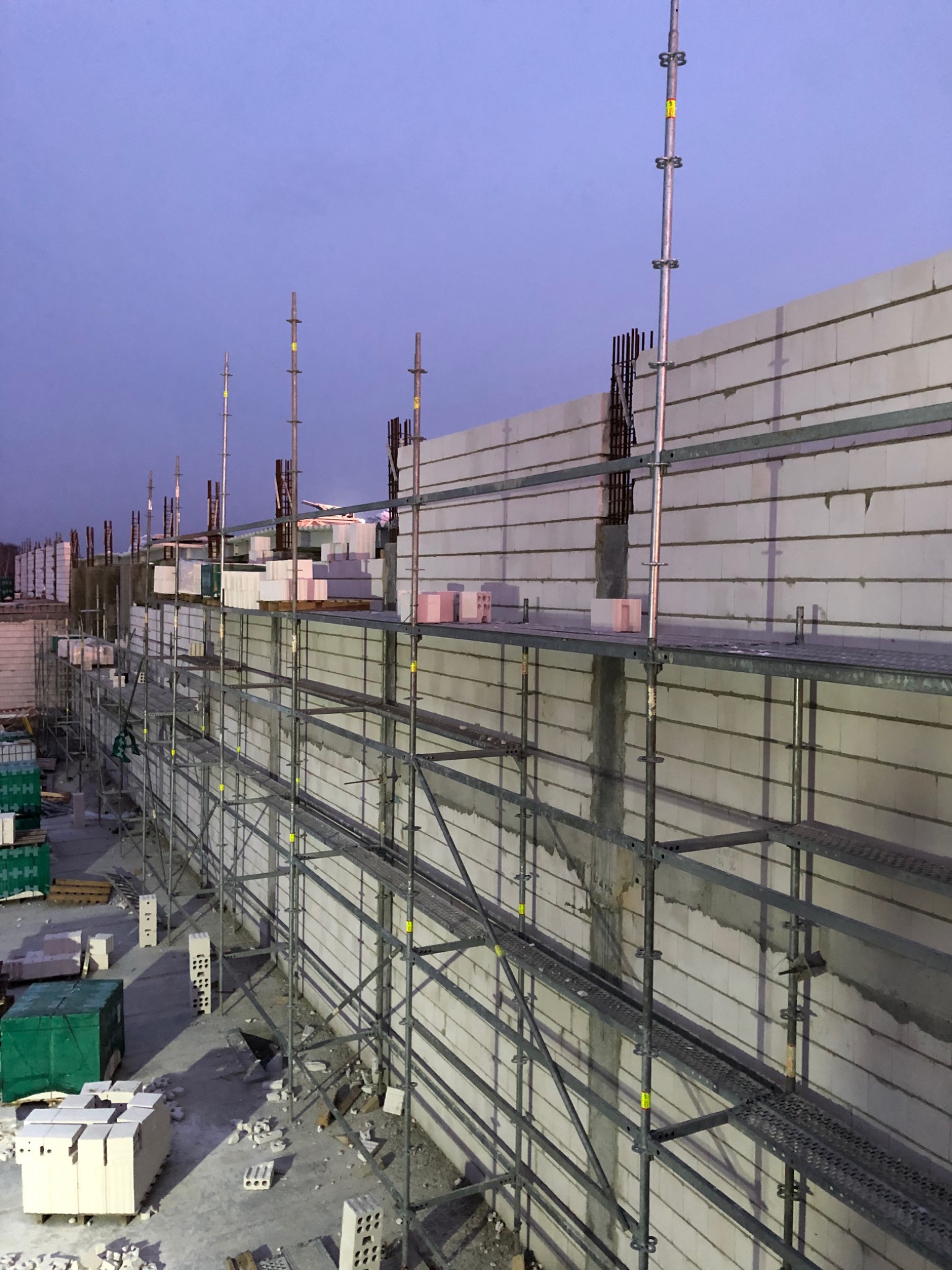 Wznoszenie ściany z betonu komórkowego Ytong na rusztowaniu, widoczne wystające pręty zbrojeniowe, zapas bloczków na paletach i resztki zaprawy na ścianie.