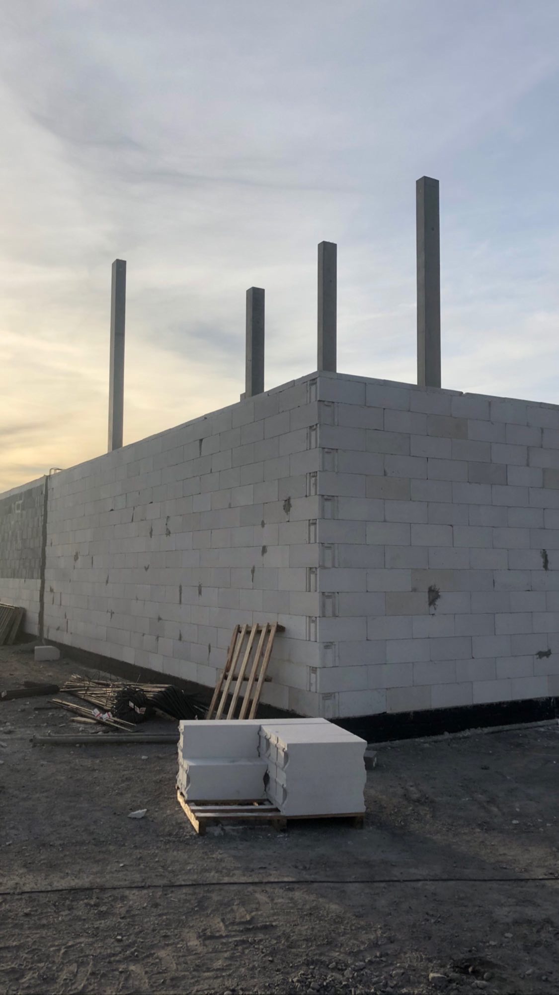 Wznoszone ściany budynku z betonu komórkowego, widoczne cztery betonowe słupy konstrukcyjne, paleta z bloczkami i materiały budowlane na placu budowy.