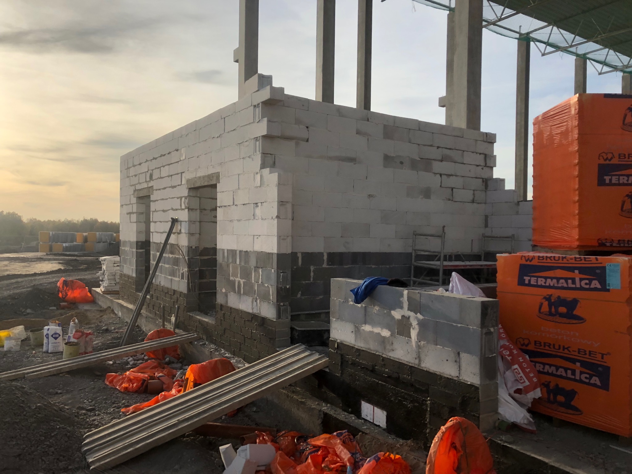 Wznoszenie ścian parteru budynku z bloczków betonu komórkowego, widoczne otwory okienne i drzwiowe, częściowo zaizolowane fundamenty, palety bloczków Termalica w pomarańczowej folii, teren budowy...
