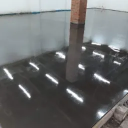 Świeżo wylana, ciemnoszara posadzka z wyraźnymi refleksami światła, częściowo otoczona drewnianym obramowaniem, z ceglanym filarem w tle.