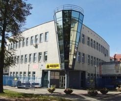Budynek szkoły lub centrum szkoleniowego z charakterystyczną, przeszkloną wieżyczką na rogu, widoczny szyld informacyjny przy wejściu.