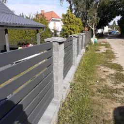 Nowoczesne ogrodzenie panelowe z szarymi przęsłami i betonowymi słupkami, widok z zewnątrz posesji, trawnik z rzadką trawą, w tle domy jednorodzinne i drzewa.