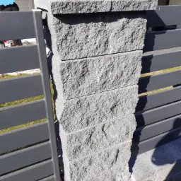 Fragment ogrodzenia z szarej, łupanej kostki kamiennej ułożonej w filar, połączonego z metalowymi przęsłami w kolorze antracytowym. Widoczne łączenie filaru z podłożem z kostki brukowej.
