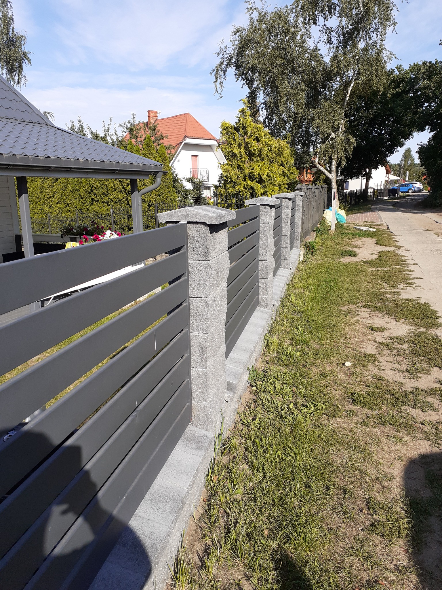 Nowoczesne ogrodzenie panelowe z szarymi przęsłami i betonowymi słupkami, widok z zewnątrz posesji, trawnik z rzadką trawą, w tle domy jednorodzinne i drzewa.