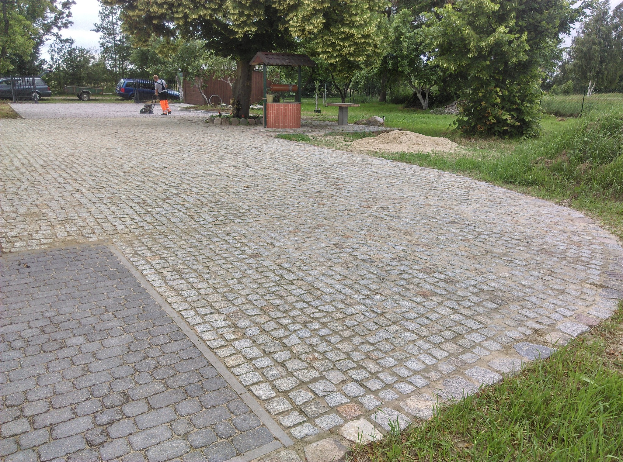Ułożona kostka brukowa z szarego granitu na dużej powierzchni placu; w tle studnia z cegły, drzewa i fragment zaparkowanych pojazdów.