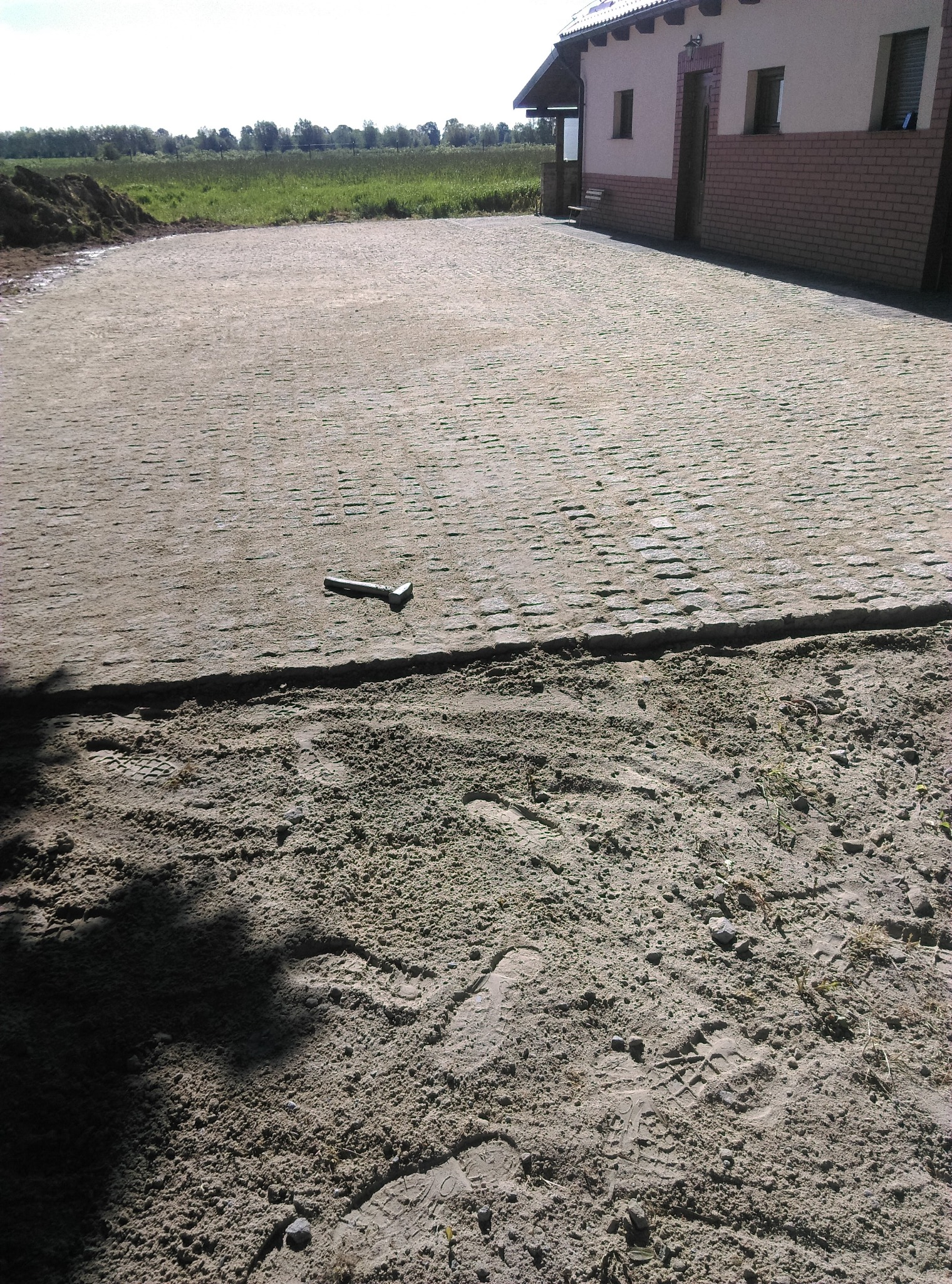 Świeżo ułożona, szara kostka brukowa na podjeździe z widocznym narzędziem do jej osadzania, obok fragment przygotowanego terenu pod dalsze prace.