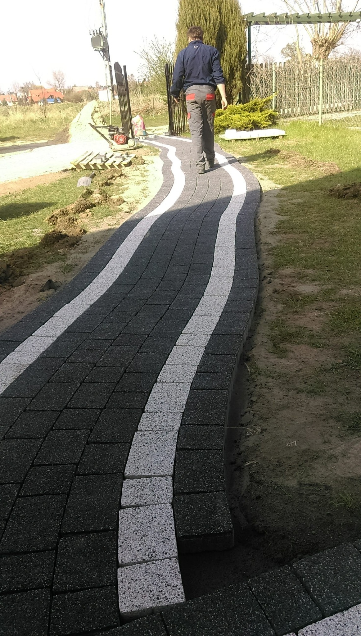 Kręta ścieżka z kostki brukowej w kolorze grafitowym z białymi pasami, prowadząca przez trawnik w kierunku furtki i spacerującego mężczyzny.