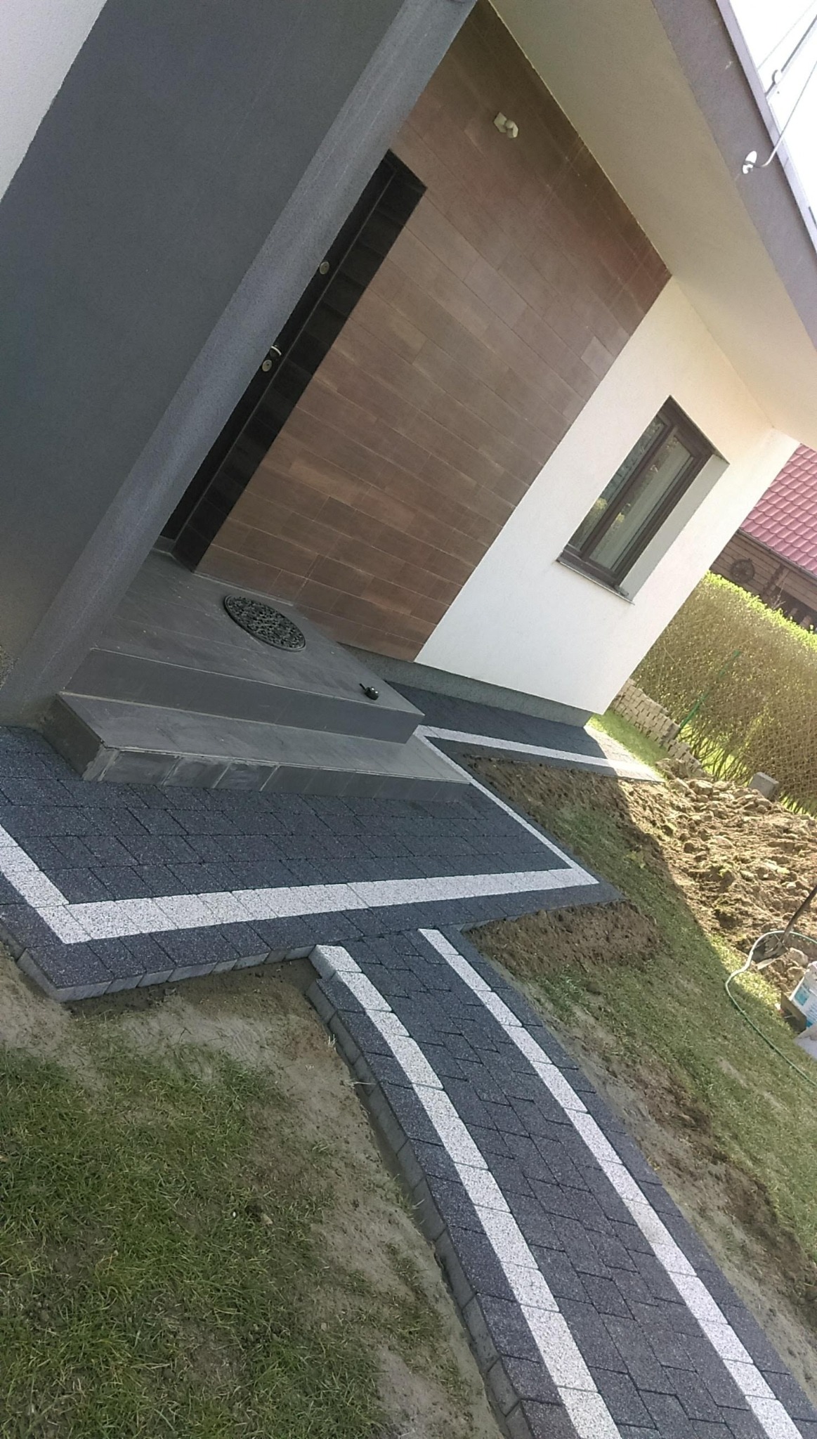 Ścieżka z ciemnej kostki brukowej z białymi paskami prowadząca do schodów wejściowych domu z brązową elewacją.