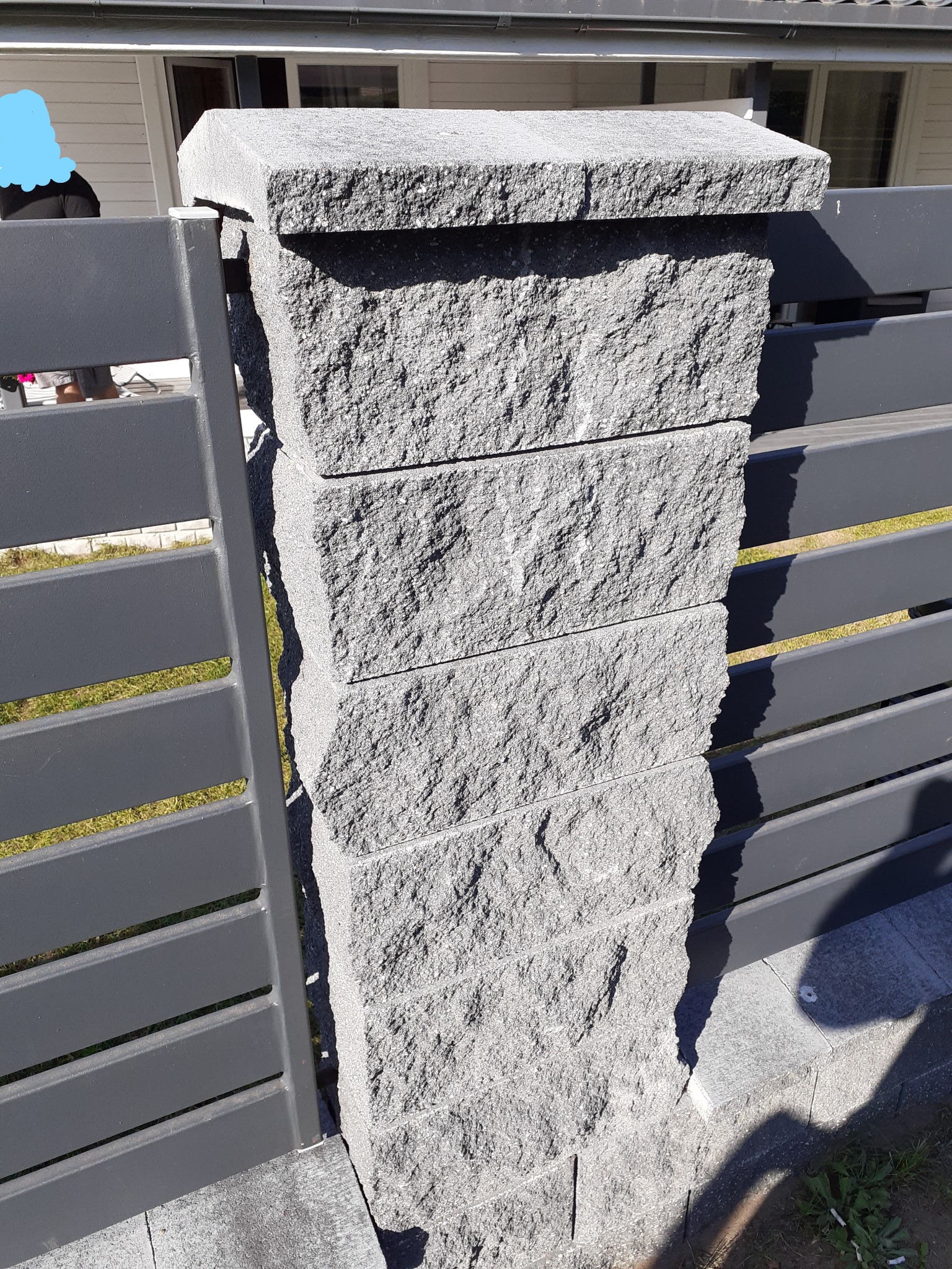 Fragment ogrodzenia z szarej, łupanej kostki kamiennej ułożonej w filar, połączonego z metalowymi przęsłami w kolorze antracytowym. Widoczne łączenie filaru z podłożem z kostki brukowej.
