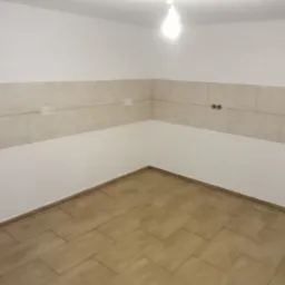 Wnętrze pomieszczenia z wyłożoną podłogą z płytek imitujących drewno i częściowo wyłożonymi płytkami ścianami, widoczne gniazdka elektryczne i otwory na kable, sufit z żar&oacute;wką.