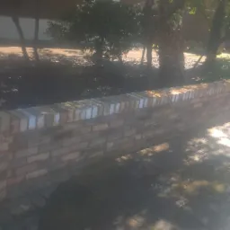 Niska, nowo wymurowana ściana z cegły klinkierowej w ogrodzie, z widocznymi fragmentami drzew i roślin w tle, oświetlona przez silne słońce.