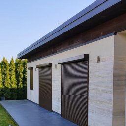 Dominika Kalinowska Rolkado - Elewacja domu z dwoma brązowymi roletami zewnętrznymi, widok z zewnątrz, z zielonym trawnikiem i tujami w tle.