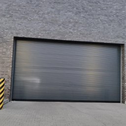 Dominika Kalinowska Rolkado - Szara, zamknięta brama rolowana w szarej ceglanej ścianie, z żółto-czarnym słupkiem ostrzegawczym po lewej stronie.