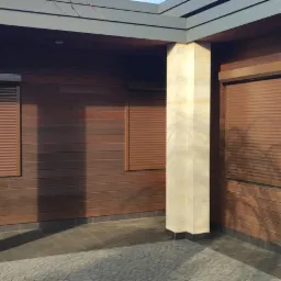 Zewnętrzne, brązowe rolety na elewacji budynku z drewnianą okładziną, widoczne kamienne filary i fragment kostki brukowej przed budynkiem.