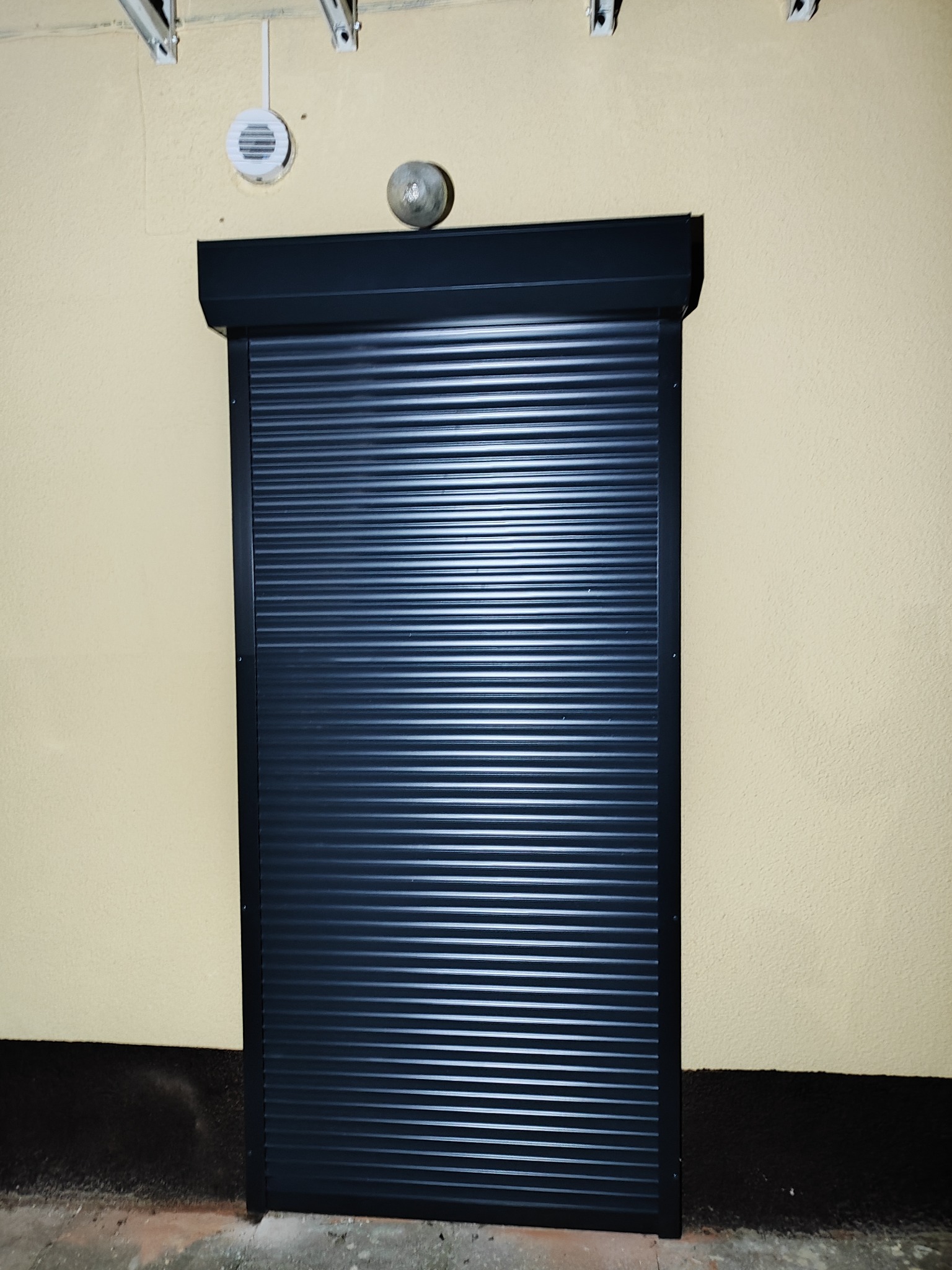 Zamknięta, ciemna roleta zewnętrzna zamontowana na beżowej ścianie budynku z widoczną wentylacją i elementami mocującymi pod sufitem.