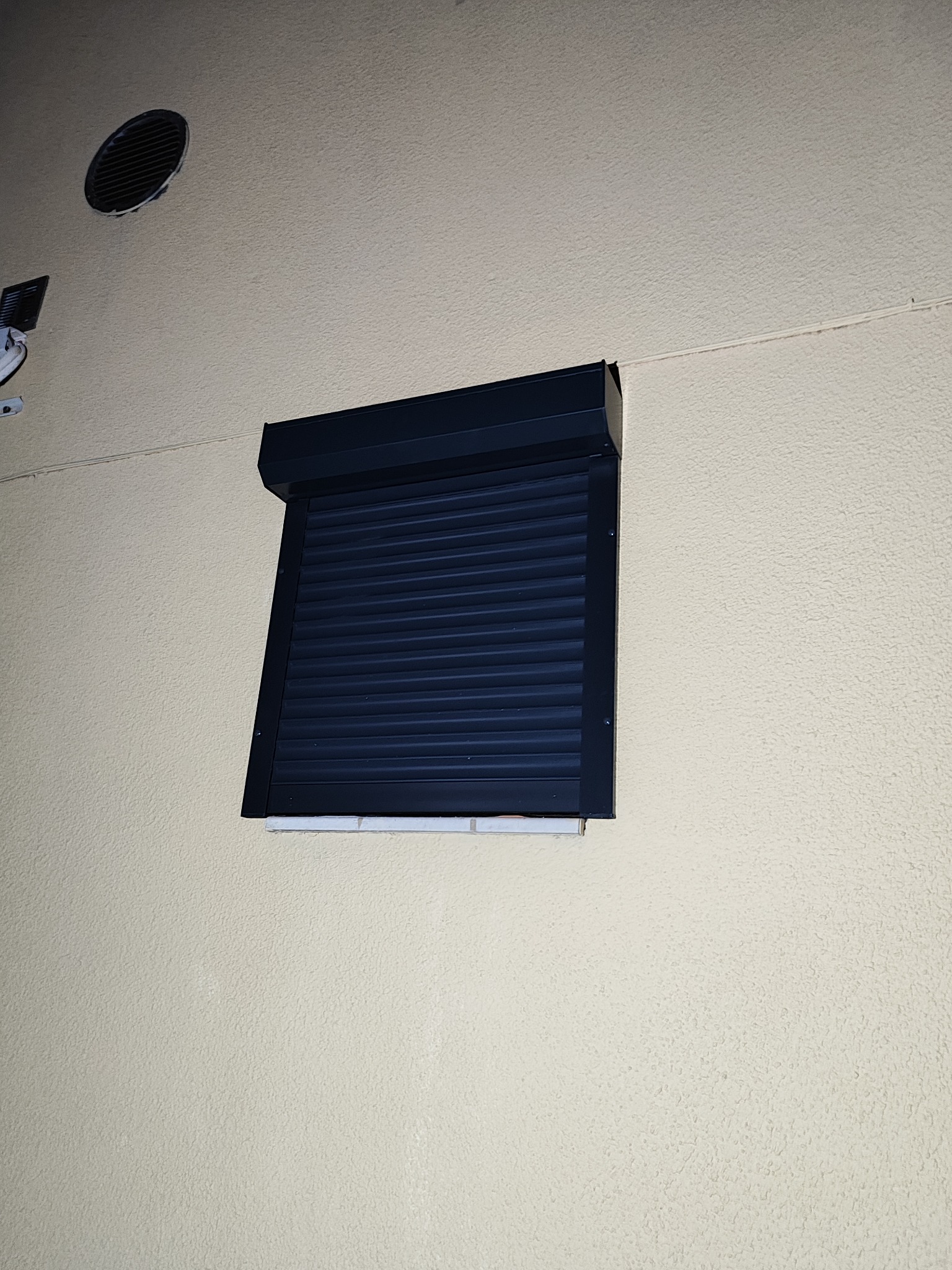 Zamknięta, ciemna roleta na elewacji budynku o jasnej, chropowatej strukturze, widoczny fragment wentylacji i kamery.