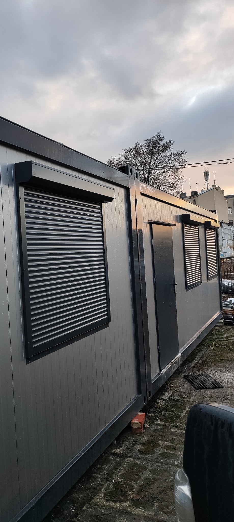 Szary kontener biurowy z zamkniętymi roletami zewnętrznymi, widoczne okna i drzwi, pochmurne niebo w tle.