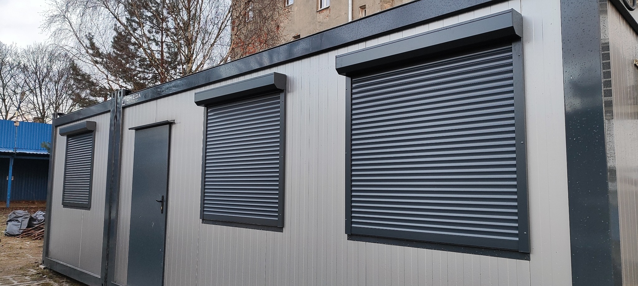 Szary kontener biurowy z trzema oknami zabezpieczonymi szarymi roletami zewnętrznymi, widok z zewnątrz w pochmurny dzień.