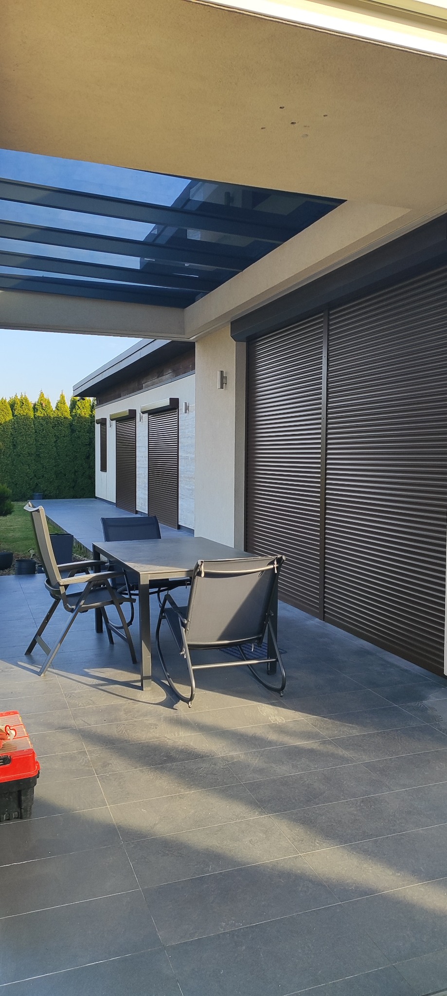 Zewnętrzne brązowe rolety na oknach domu z tarasem, na którym stoi szary stół i krzesła; skrzynka z narzędziami widoczna w rogu kadru.