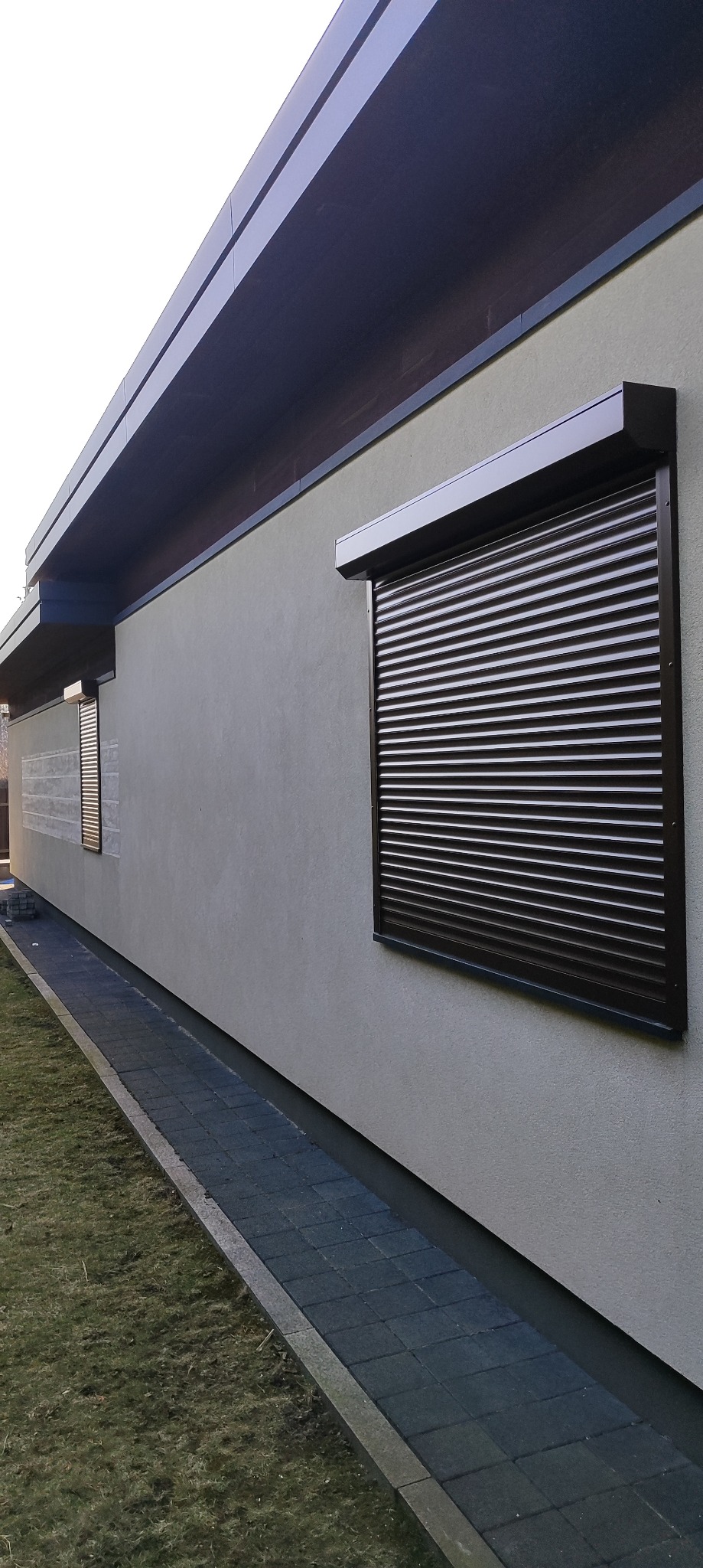Zewnętrzna roleta w kolorze ciemnego brązu, zamontowana na jasnej elewacji domu, widok z boku z perspektywą na trawnik i chodnik.