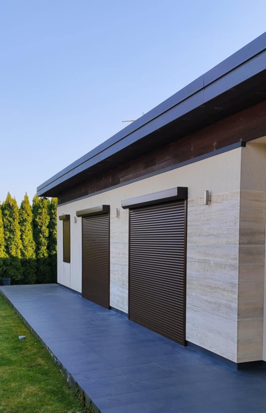 Elewacja domu z dwoma brązowymi roletami zewnętrznymi, widok z zewnątrz, z zielonym trawnikiem i tujami w tle.