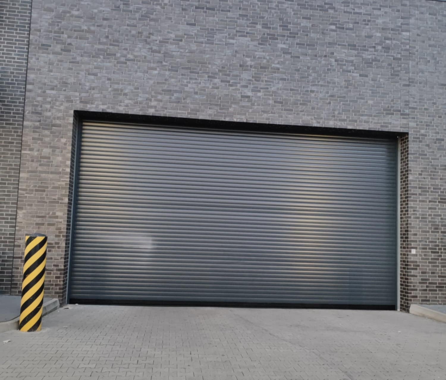 Szara, zamknięta brama rolowana w szarej ceglanej ścianie, z żółto-czarnym słupkiem ostrzegawczym po lewej stronie.
